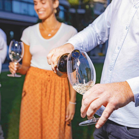 Person pouring wine into a glass with a blurred background of people and greenery